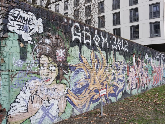 The colourful breeze block wall at the East Side Gallery, 2 February 2022 The graffiti covered breeze block wall. Over the back end of the wall, a newly constructed building overhangs.