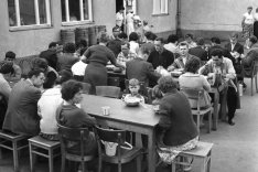 Drei lange Tische stehen hintereinander in einem Hof. An ihnen sitzen essende Frauen, Männer und Kinder.
