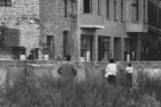 Ein Mann, eine Frau und ein Kind schauen über die Mauer, sie sind von hinten zu sehen. Auf der anderen Seite der Mauer steht eine Häuserzeile. Im zweiten Stock eines Wohnhauses stehen Menschen am Fenster.