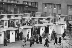 Vor dem Bahnhof stehen in mehreren Reihen zwanzig kleine Häuschen, darin und darum sind viele Menschen versammelt.