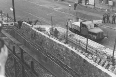 Unscharf im Vordergrund am linken Bildrand ist eine Hand mit Fotokamera zu erkennen. Quer durch das Bild zieht sich die Mauer aus Hohlblocksteinen, die gerade aufgebaut wird. Dahinter sieht man Arbeiter, eine Straßensperre aus Betonelementen, einen Lastwagen, drei Polizisten und zwei Passanten.